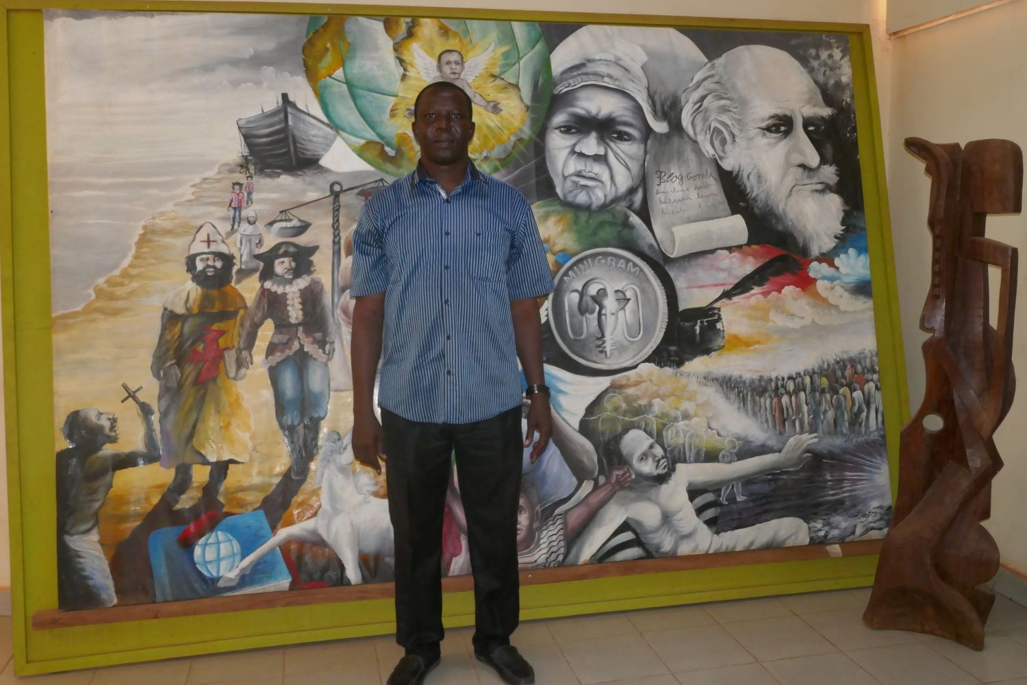 Sinaly DJIBO, directeur des expositions et de la médiation du musée national, pose avec un énorme tableau réalisé par un jeune artiste en résidence. Le tableau évoque la colonisation et l’esclavage, l’indépendance et l’importance des jeunes générations pour le développement du pays © Globe Reporters