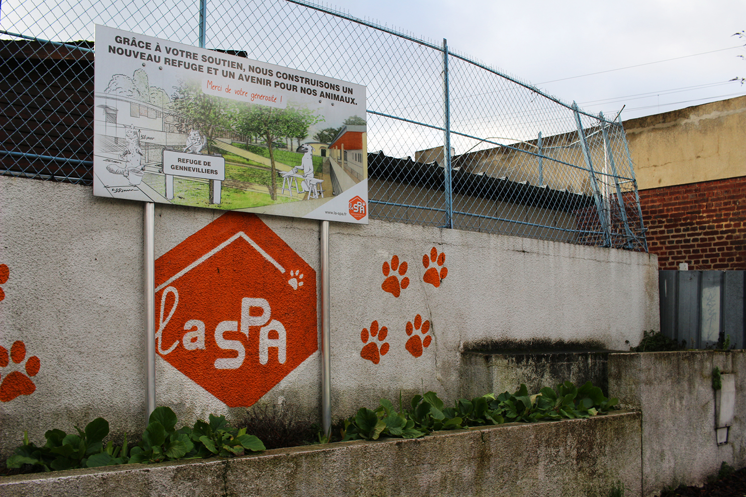 La SPA la plus proche de Paris, qui aussi la plus ancienne, se situe à quelques mètres de la gare RER de Gennevilliers. Le refuge se trouve derrière ces hauts murs et une grande porte coulissante en tôle à proximité de la route. Sur le panneau, nous pouvons apercevoir un dessin qui donne quelques informations sur le futur nouveau refuge pour remplacer celui-ci © Globe Reporters