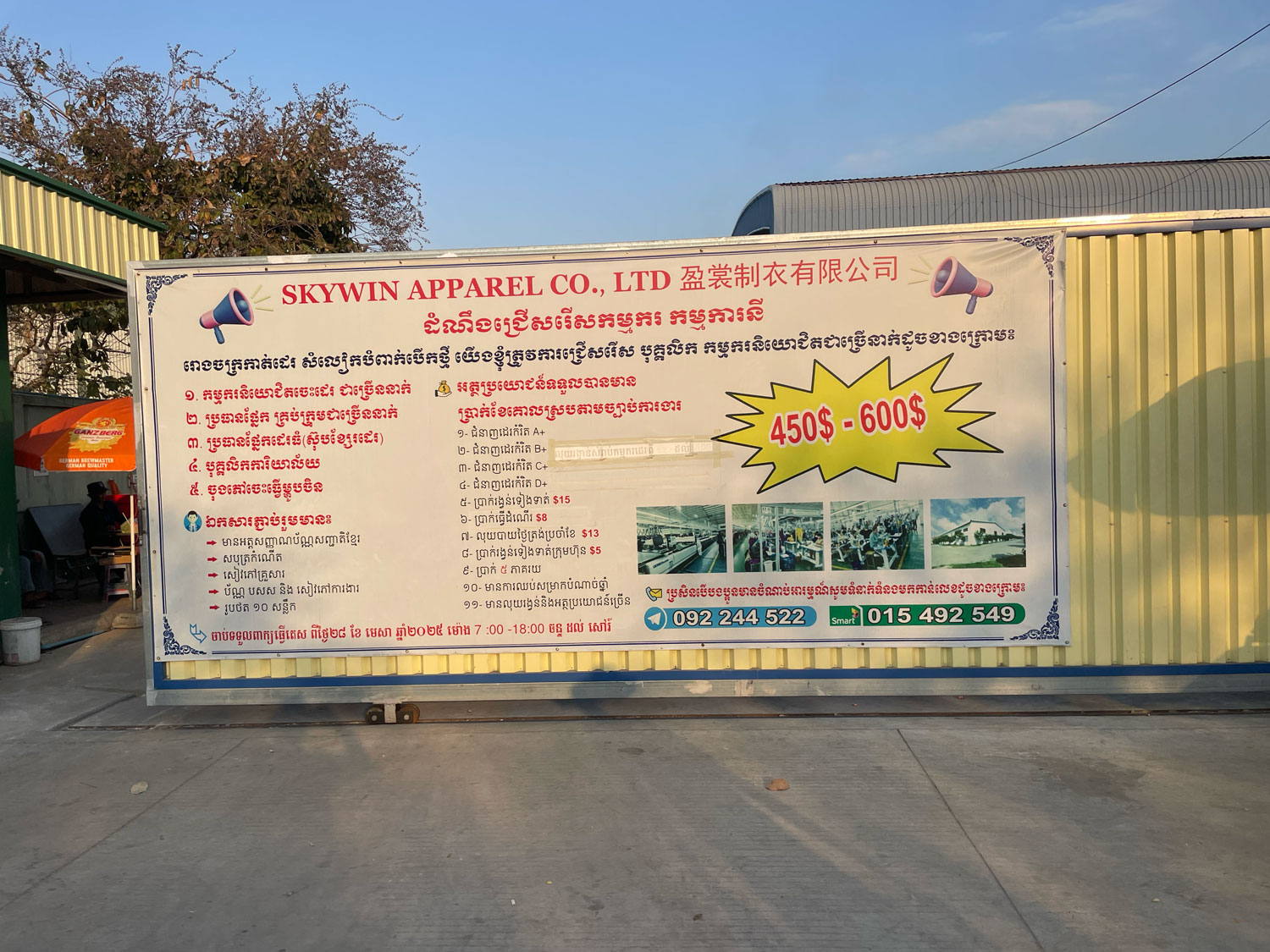 Sur le portail de l’usine Skywin Apparel Co. une affiche annonce un recrutement urgent de 450 à 600 employés (couturiers, opérateurs de machines à coudre, etc.) avec un salaire attractif mis en avant. L’usine produit du prêt à porter pour les marques Disney, Marvel, HEAD, Tony Hawk, Proplayer, Route 66, Bonjour, Fila et Championship Gold © Globe Reporters